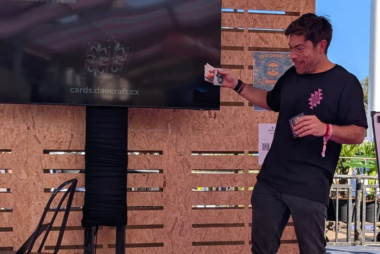 A facilitator holds up cards in front of a screen that shows the deck and cards.daocraft.cx.